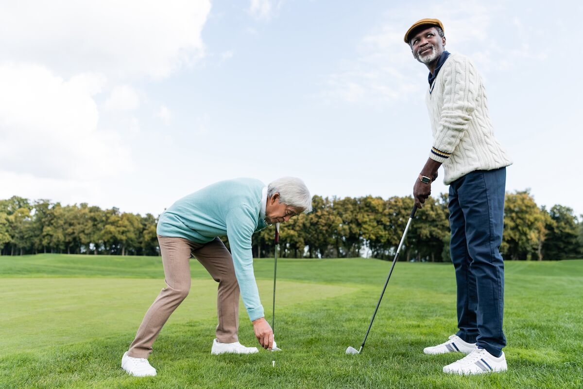 asian senior man putting ball on golf tee near african american friend