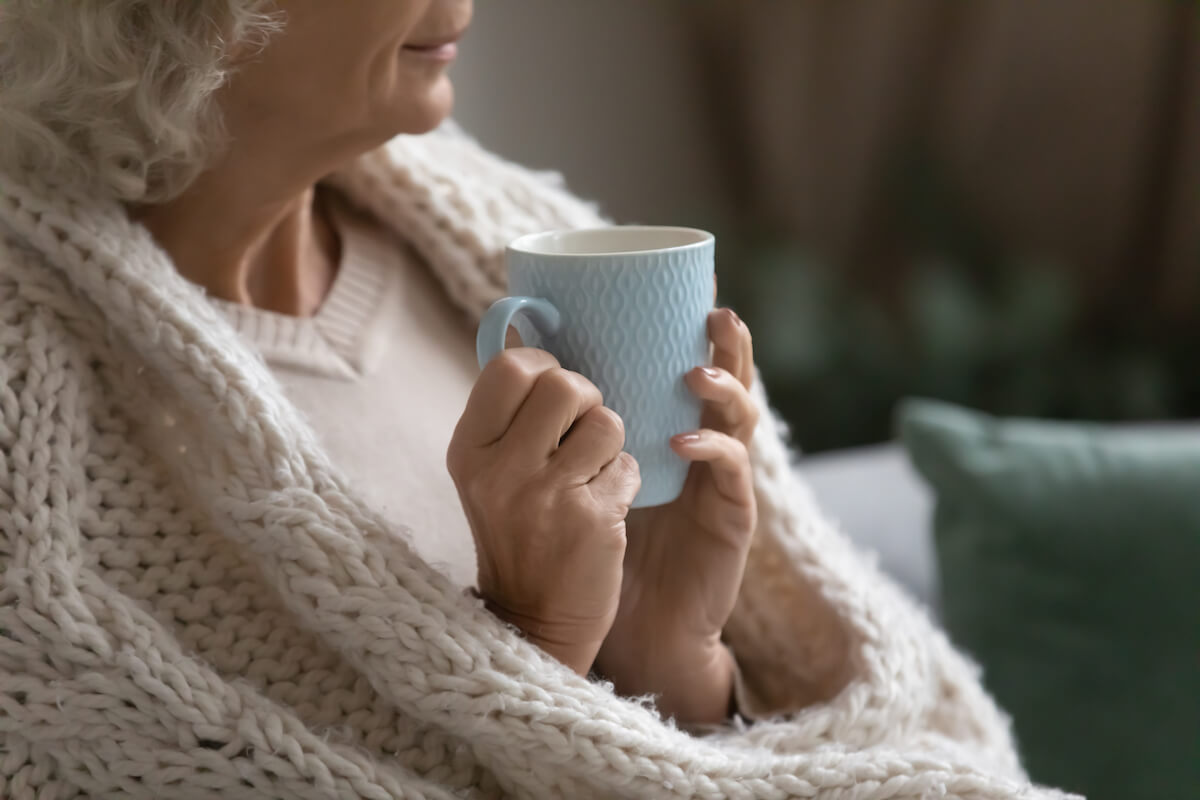 Aged calm woman resting wrapped in knitted plaid drink evening tea look into distance, close up.